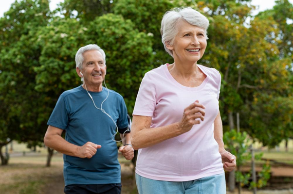 Um casal de idosos corre alegremente em um parque, simbolizando o conceito de "envelhecer com saúde". A mulher, com cabelo curto e grisalho, veste uma camiseta rosa, enquanto o homem, com cabelo branco, usa uma camiseta azul. Ambos exibem sorrisos e parecem estar se divertindo enquanto se exercitam ao ar livre, cercados por árvores verdes e um ambiente natural.