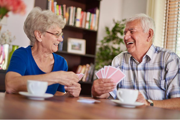 Um casal de idosos está sentado à mesa, sorrindo e se divertindo enquanto jogam cartas. Ambos têm xícaras de café à sua frente. A mulher, com cabelo curto e grisalho, veste uma blusa azul, enquanto o homem, com cabelo branco, usa uma camisa xadrez. O ambiente é acolhedor, com prateleiras de livros ao fundo e luz natural entrando pela janela.