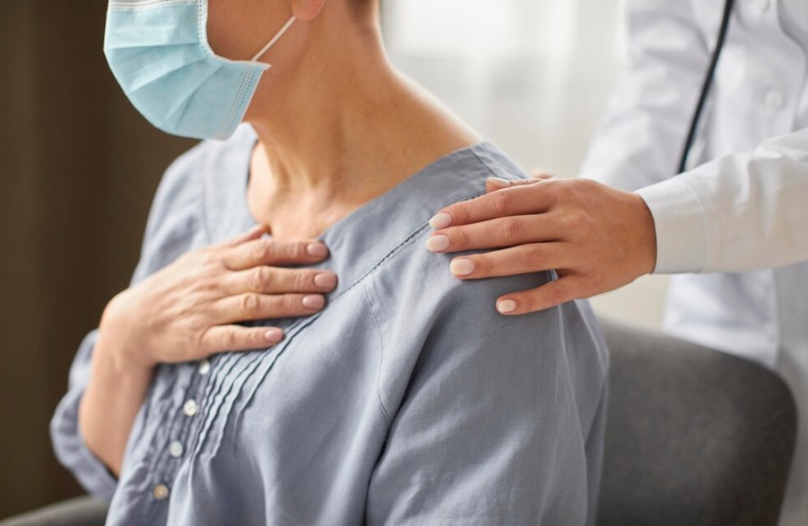 Mulher em consulta médica, com máscara, demonstrando preocupação ao tocar o peito, enquanto um profissional de saúde a conforta