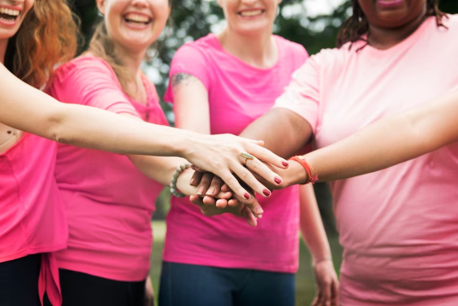 Grupo de mulheres sorrindo e unindo as mãos em um gesto de apoio, vestindo camisetas rosas, simbolizando a campanha Outubro Rosa.