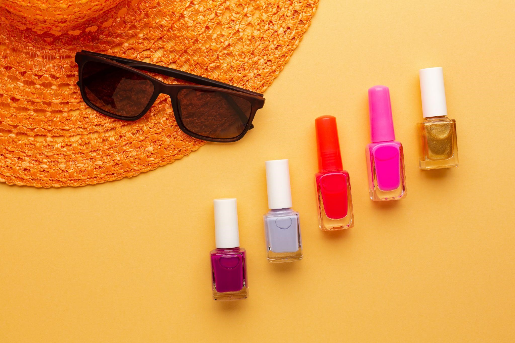 Frascos de esmaltes com cores vibrantes ao lado de uma chapéu de praia e óculos de sol, simbolizando as cores de esmaltes tendências para o verão.